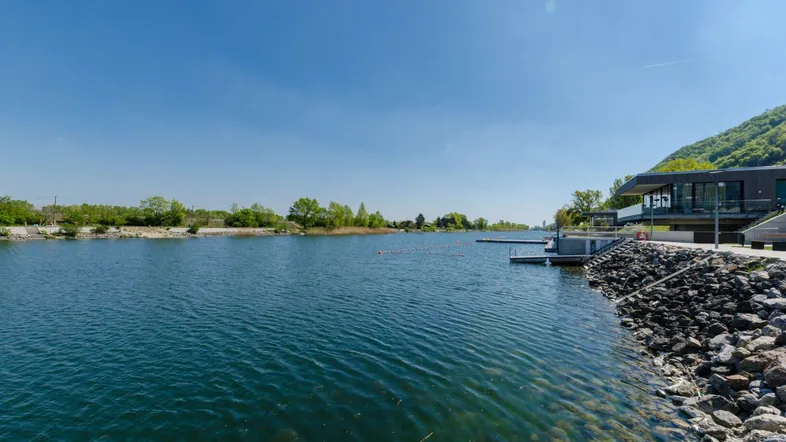 Weitläufiger See mit Uferbebauung und grünen Hügeln im Hintergrund, ideal für Wassersport.