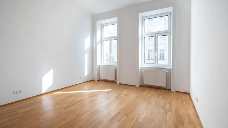 Helles Zimmer mit Parkettboden und zwei großen Fenstern, das viel Tageslicht hereinlässt.