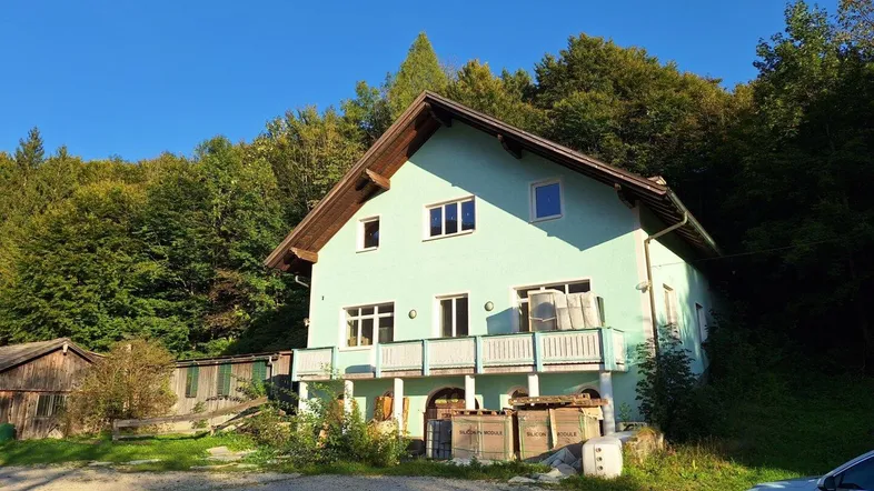 Grünes Haus mit Holzbalkonen und Satteldach, umgeben von Bäumen in ländlicher Umgebung.