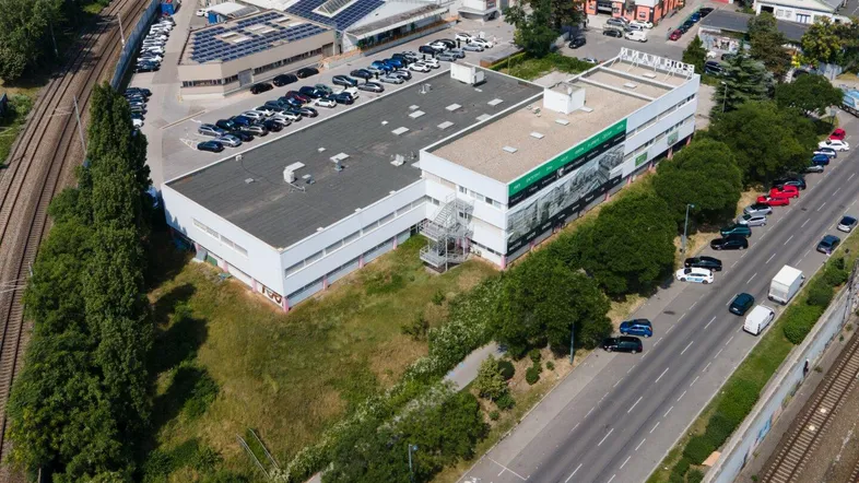 Luftaufnahme eines Gewerbekomplexes mit Parkplätzen, Hauptstraße und Bahngleisen in sonniger Umgebung.
