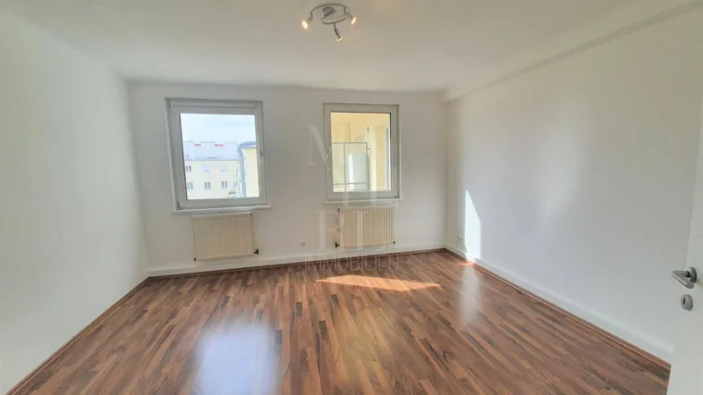 Helles Zimmer mit Parkettboden, weißen Wänden und zwei Fenstern, in gutem Zustand.
