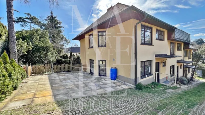 Gelbe Fassade des Hauses mit gepflasterter Einfahrt und Gartenbereich unter blauem Himmel.