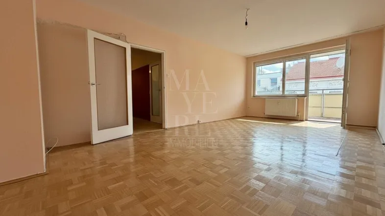 Geräumiges Wohnzimmer mit Fischgrätparkettboden, hellen Wänden und Zugang zum Balkon.