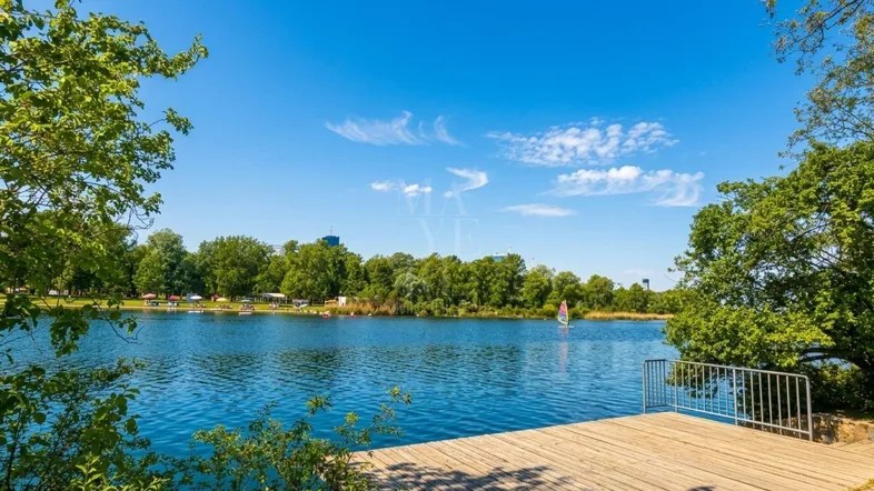 Idyllischer See mit Holzsteg und üppiger grüner Uferlandschaft unter strahlend blauem Himmel.