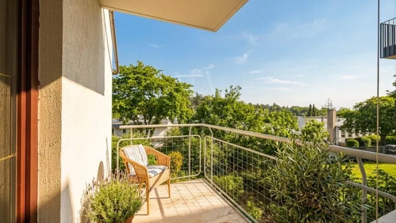 Sonniger Balkon mit Rattan-Sessel, Pflanzen und weitem Blick ins Grüne.