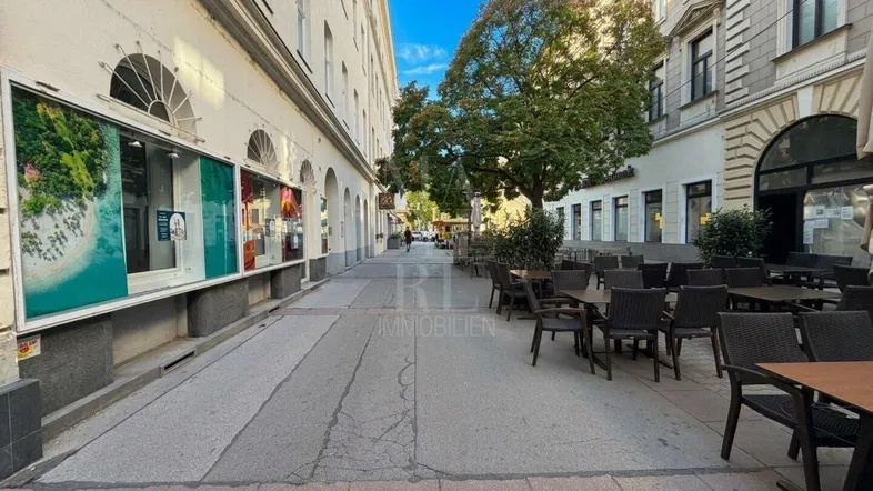 Eine belebte Straße mit Gebäuden, Geschäften und einem Café mit Außensitzplätzen unter Bäumen.