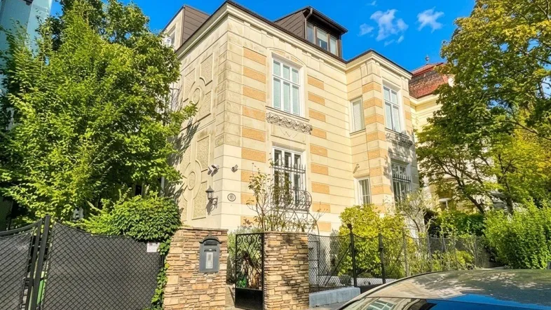 Klassisches Gebäude mit heller, gestreifter Fassade, verzierten Fenstern und üppiger Begrünung unter blauem Himmel.