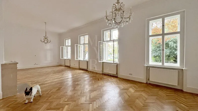 Großzügiges Wohnzimmer mit Fischgrätparkett, hohen Fenstern und eleganten Kronleuchtern, das viel Licht hereinlässt.
