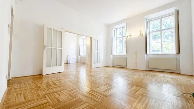 Geräumiges Wohnzimmer mit Fischgrätparkettboden, hohen Decken und großen Fenstern, die viel Tageslicht hereinlassen.