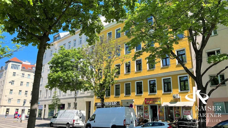 Gelbes Mehrfamilienhaus an einer belebten Straße mit Bäumen und Geschäften im Erdgeschoss.