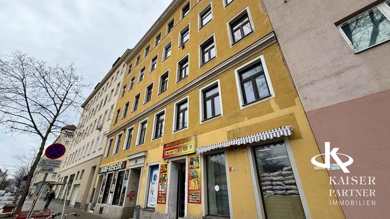 Mehrstöckiges Wohn- und Geschäftshaus mit gelber Fassade und Ladenlokalen im Erdgeschoss an einer Stadtstraße.