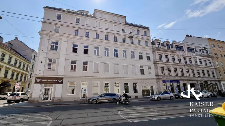 Fassade eines Mehrfamilienhauses an einer belebten Straße mit Geschäften im Erdgeschoss und vielen Fenstern.
