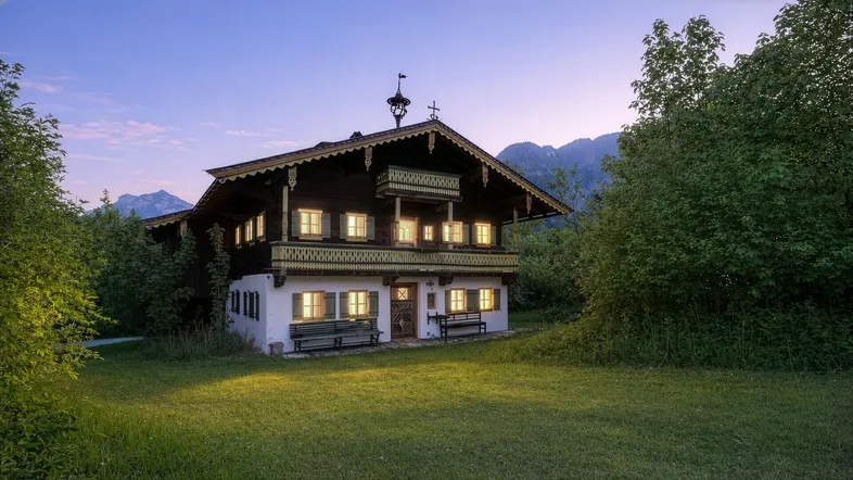 Traditionelles Bauernhaus im alpenländischen Stil bei Sonnenuntergang mit beleuchteten Fenstern und Bergblick.