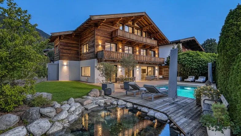 Luxuriöses Chalet im alpinen Stil mit beleuchtetem Garten, Pool und Teich bei Abenddämmerung.