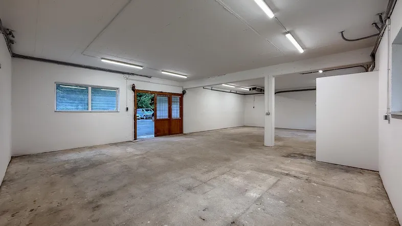 Großer, leerer Raum mit Betonboden, weißen Wänden und einer breiten Holztür, ideal für Lager oder Werkstatt.