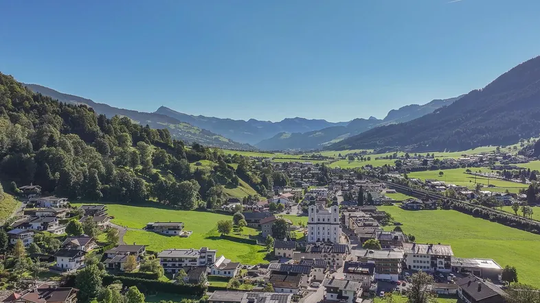 Panoramablick auf ein malerisches Dorf in einem grünen Tal, umgeben von Bergen unter blauem Himmel.