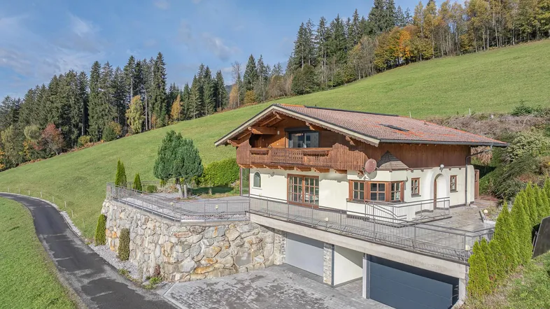 Charmantes Haus im alpinen Stil mit großer Terrasse und Doppelgarage, eingebettet in eine grüne Hügellandschaft.