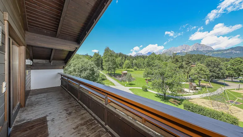 Geräumiger Holzbalkon mit Blick auf einen Spielplatz und die majestätische Bergwelt.