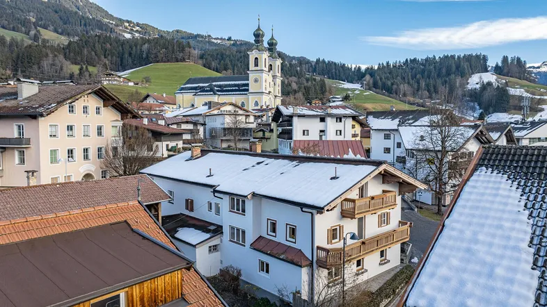 Luftaufnahme eines verschneiten Dorfes mit Kirche und Bergen im Hintergrund.