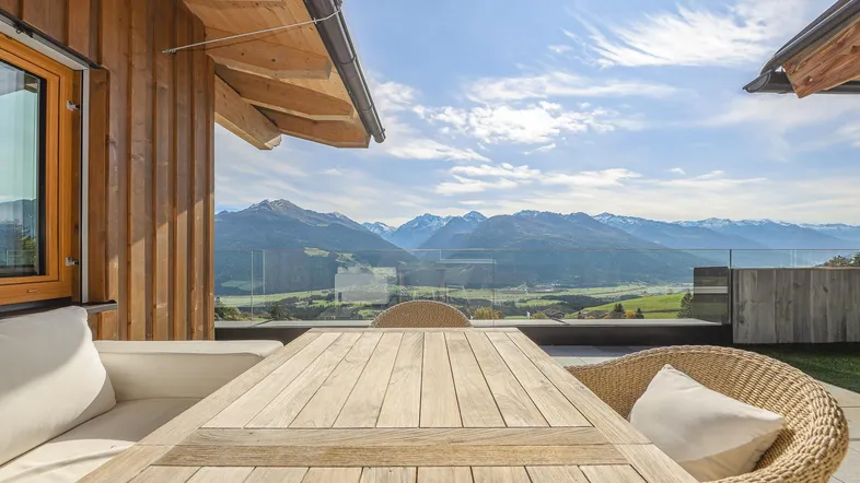 Große Terrasse mit Holztisch, Korbsesseln und atemberaubendem Bergblick unter blauem Himmel.