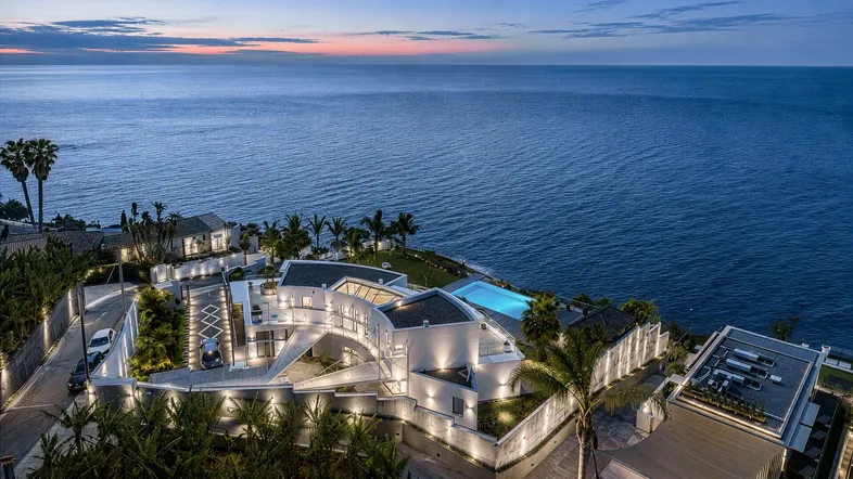 Moderne Villa mit beleuchtetem Außenbereich und Infinity-Pool bei Sonnenuntergang, direkt am Meer gelegen.