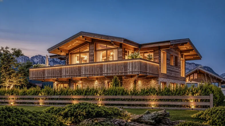 Luxuriöses Holzchalet mit Steinverkleidung und beleuchtetem Balkon vor alpiner Bergkulisse bei Dämmerung.