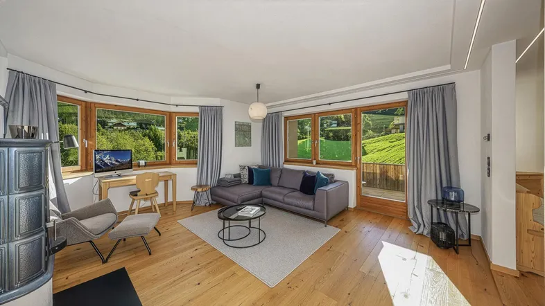 Helles Wohnzimmer mit gemütlichem Ecksofa, Kachelofen und Panoramablick auf die grüne Landschaft.