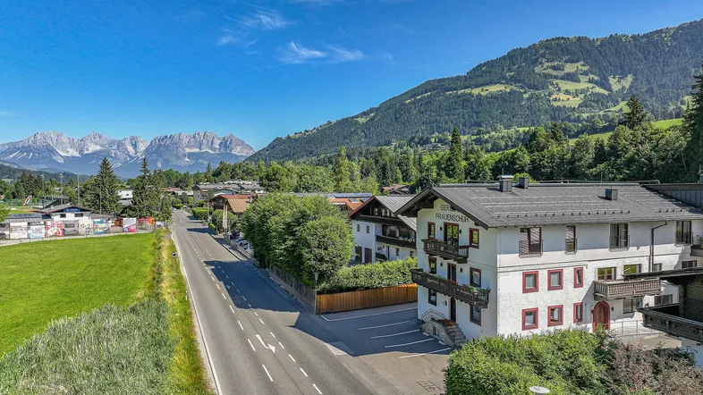 Gebäudeansicht mit traditioneller Architektur, umgeben von grünen Wiesen und majestätischen Bergen unter blauem Himmel.