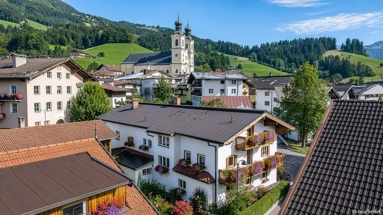 Malerische Luftaufnahme eines Bergdorfes mit Kirche und traditionellen Häusern vor grüner Bergkulisse.