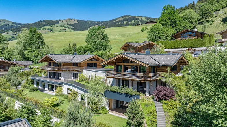 Luxuriöse Chalets in alpiner Landschaft mit grünen Hügeln und bewaldeten Bergen im Hintergrund.