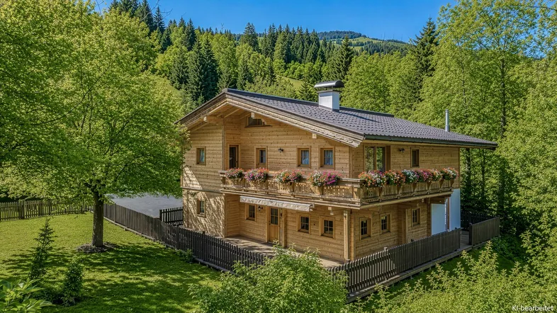 Traditionelles Holzhaus im alpinen Stil mit Blumenkästen und Bergblick, umgeben von grüner Natur.