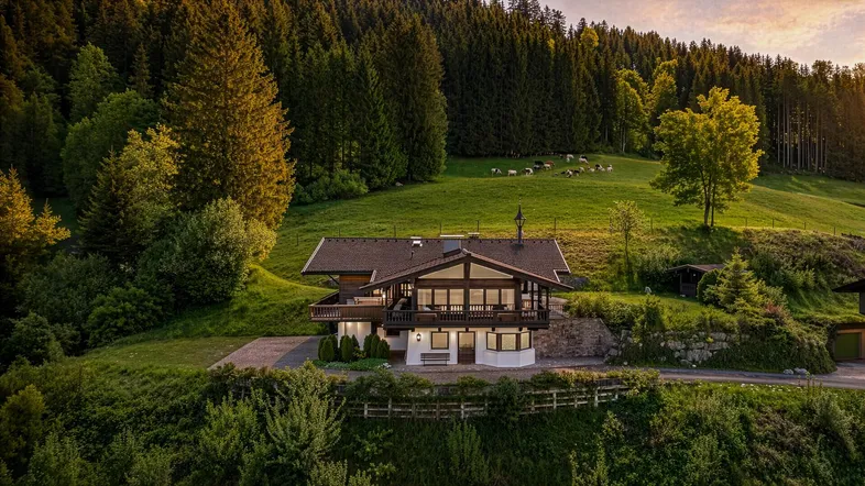Haus im alpenländischen Stil mit Bergblick, umgeben von grünen Hügeln und Wäldern bei Sonnenuntergang.