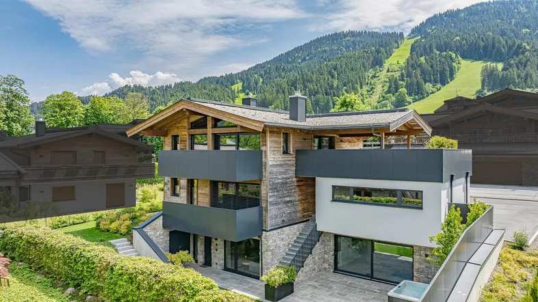 Modernes Chalet mit Holzfassade, Balkonen und Bergblick vor einer grünen Berglandschaft.