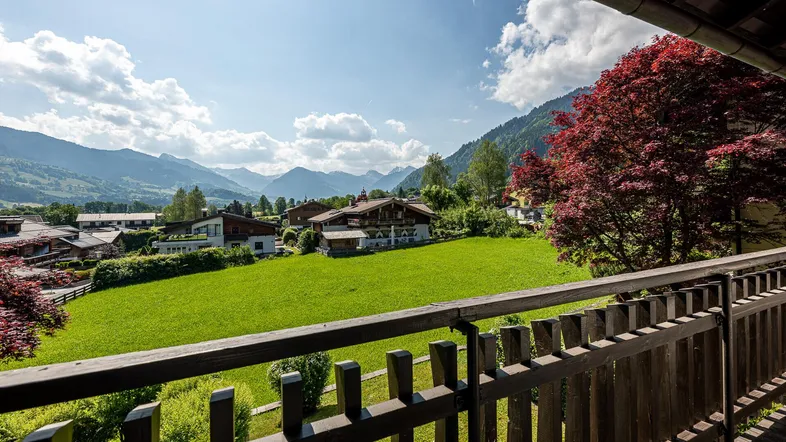 Panoramablick vom Balkon über grüne Wiesen, Häuser und die umliegende Berglandschaft an einem sonnigen Tag.