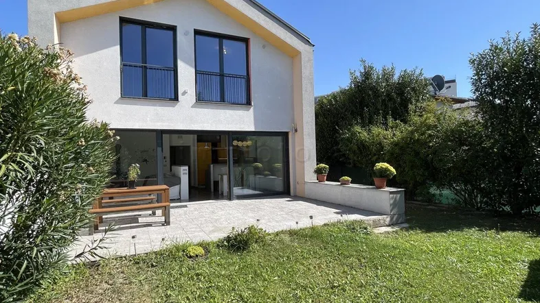 Moderne Hausfassade mit großer Terrasse und gepflegtem Garten unter blauem Himmel.