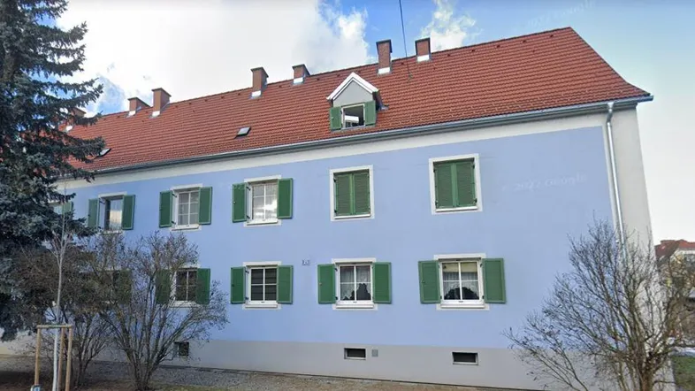 Blaues Mehrfamilienhaus mit rotem Ziegeldach, grünen Fensterläden und Bäumen im Vordergrund.