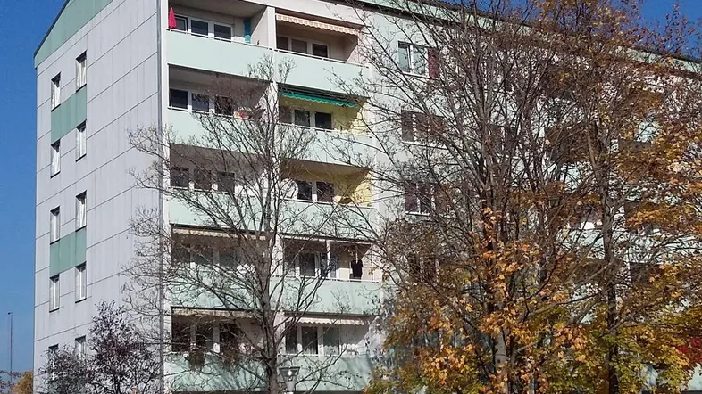 Mehrstöckiges Wohngebäude mit Balkonen und Bäumen im Herbstlaub unter blauem Himmel.