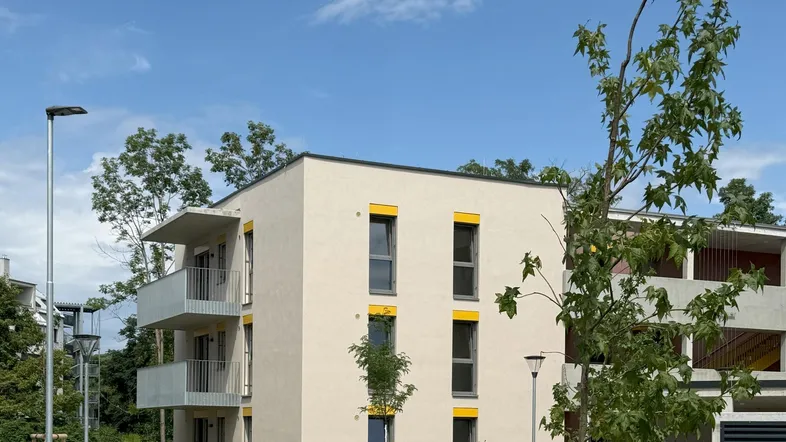 Modernes Mehrfamilienhaus mit hellbeiger Fassade, gelben Akzenten und Balkonen unter blauem Himmel.