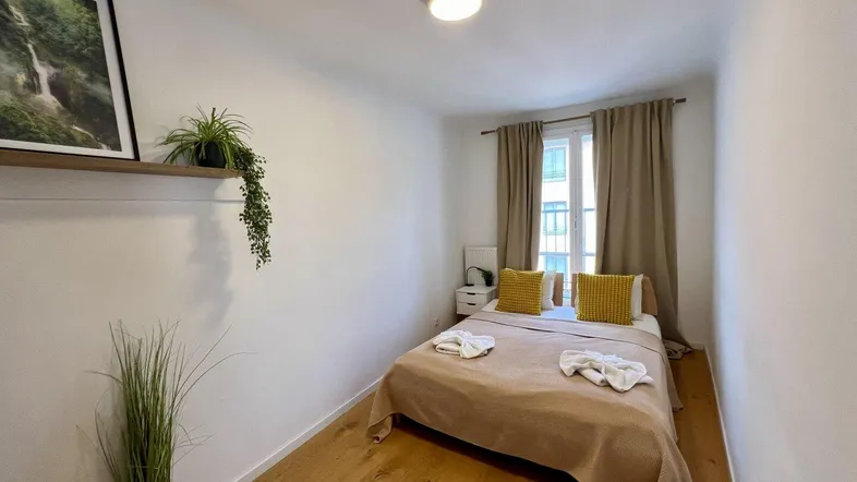 Helles Schlafzimmer mit Holzboden, beigem Bett und einem kleinen französischen Balkon.