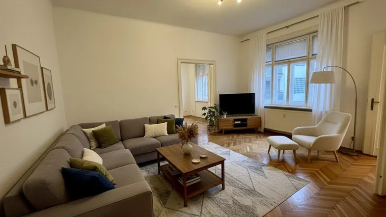 Gemütliches Wohnzimmer mit großem Ecksofa, Holzboden, hohen Decken und Blick auf die Straße.