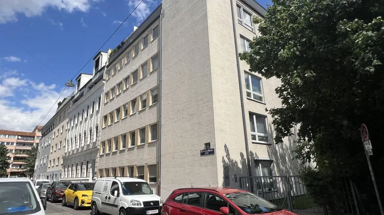 Ansicht der Gebäudefassade von der Straße mit parkenden Autos und Bäumen.
