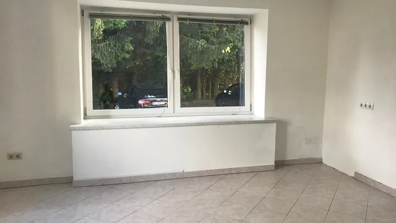 Heller Büroraum mit Fliesenboden und großem Fenster, das einen Blick ins Grüne bietet.