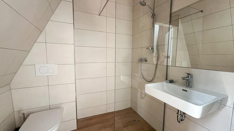 Modernes Badezimmer mit begehbarer Dusche, WC, Waschbecken und Dachschräge, hell gefliest.