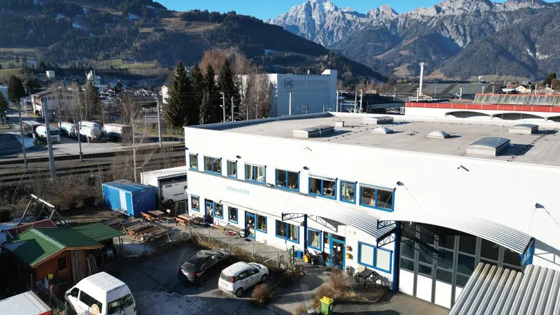 Luftaufnahme eines modernen Gebäudes mit Parkplätzen und einer beeindruckenden Bergkulisse unter strahlend blauem Himmel.