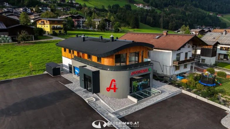Modernes Geschäftsgebäude mit Apotheke im Erdgeschoss und Wohnungen darüber, umgeben von grüner Landschaft.