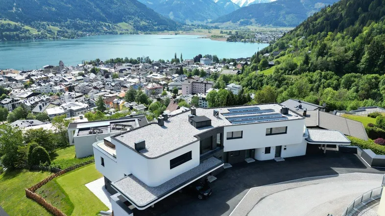 Drohnenaufnahme eines modernen Gebäudekomplexes mit Seeblick und Bergpanorama.