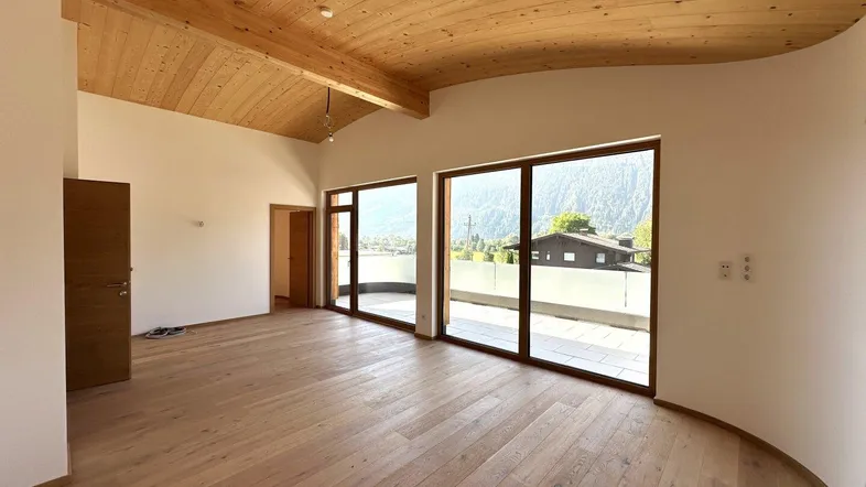 Heller Wohnbereich mit Holzboden und Holzdecke, großen Fenstern und Zugang zur Terrasse mit Bergblick.