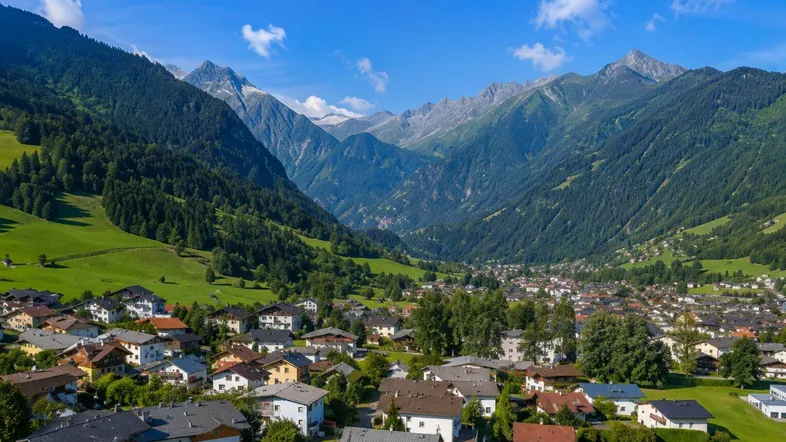 Luftaufnahme eines malerischen Dorfes in einem grünen Tal, umgeben von majestätischen Bergen unter blauem Himmel.
