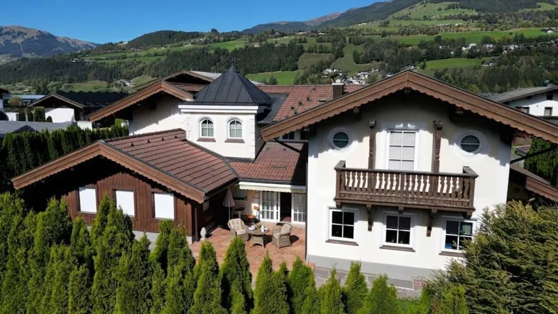Traditionelles Haus mit Satteldach, Balkon und gepflegter Hecke, umgeben von grünen Bergen.