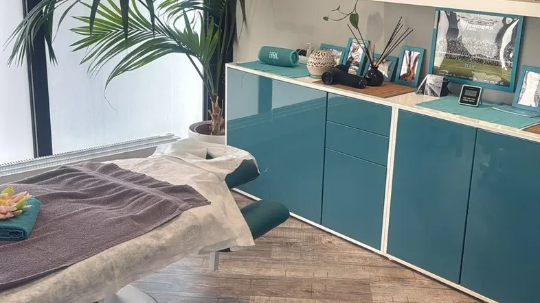 Behandlungsraum mit Massageliege, Holzfußboden und modernen blauen Schränken, gut beleuchtet.